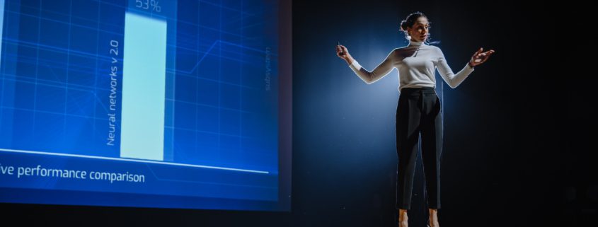 woman standing on stage with a blue monochrome graph projection behind her as she speaks